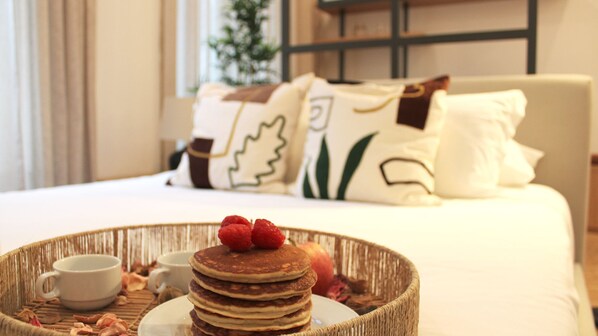 A breakfast tray with pancakes, strawberries, and a cup on a woven mat, with a bed and pillows in the background.