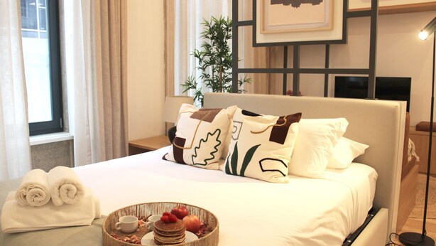 A hotel room with a neatly made bed, a bedside table with a lamp, a window with curtains, and a tray with breakfast items. - Modern Flat Near Trindade by LovelyStay (Porto)