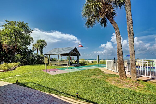 A gazebo with an American flag, surrounded by grass and palm trees, with a pool in the background.