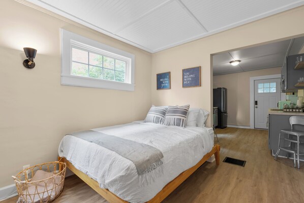 A bedroom with a bed, a window, a wall-mounted light, and a kitchen area visible through an open door. - Carriage Cottage- in the Heart of Everything Delaware (Delaware)