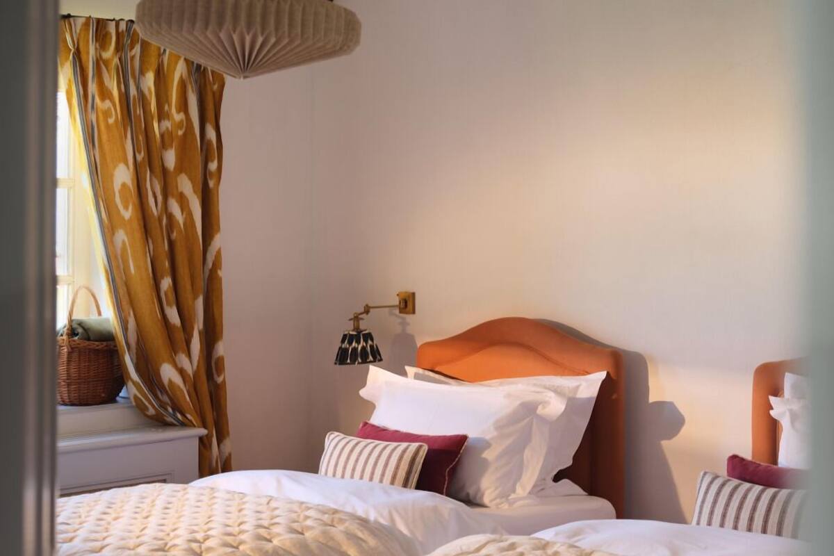 Um quarto aconchegante com duas camas, um lustre pendente e uma janela com cortinas.