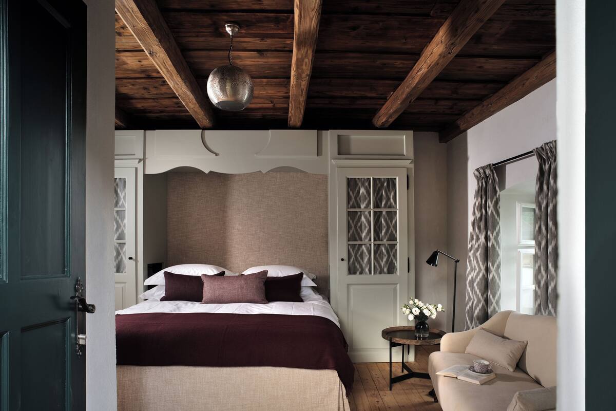 Quarto com teto de madeira, cama com cabeceira, mesa de cabeceira com abajur e uma cadeira.