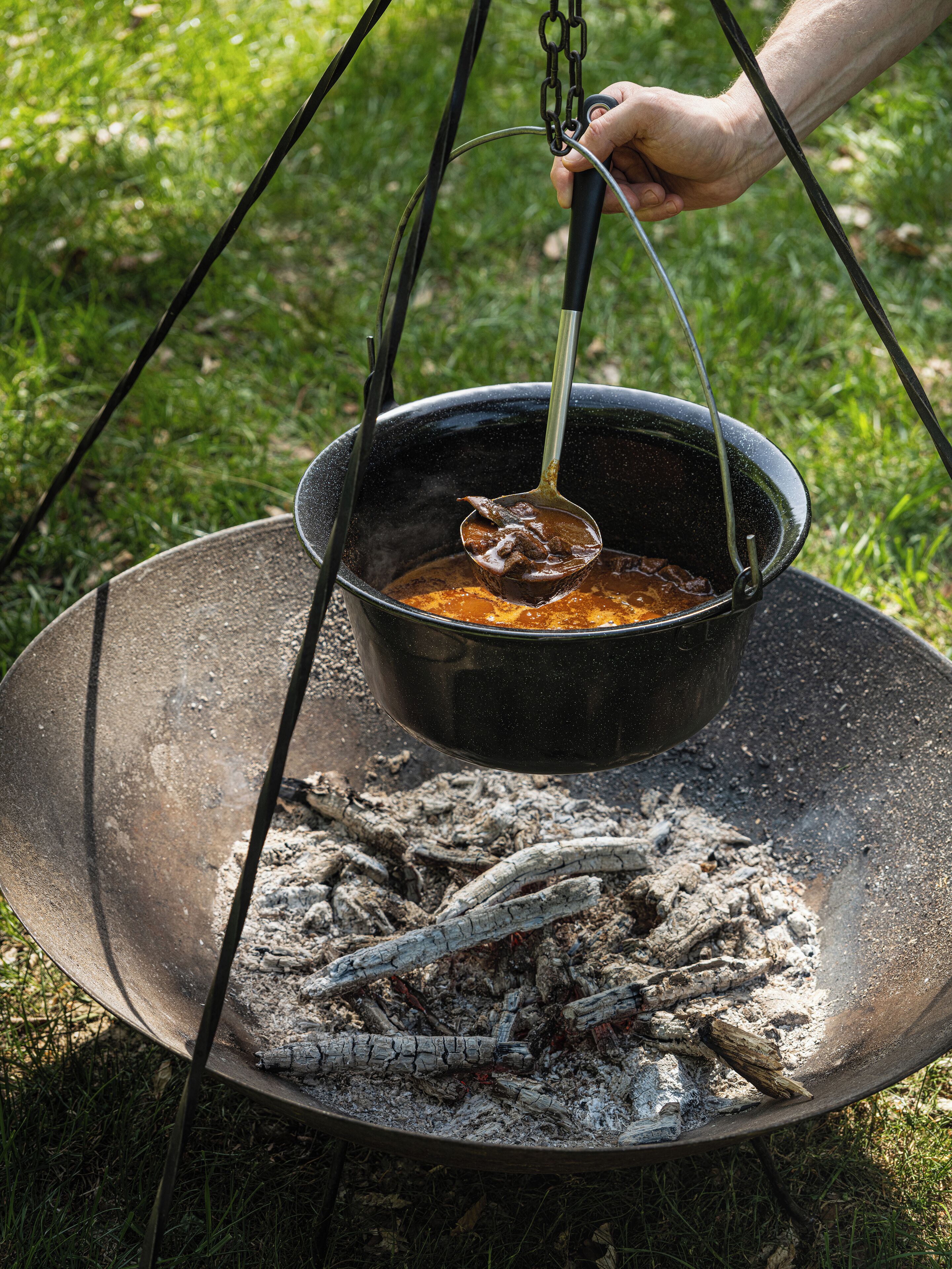 A black pot over an open fire with a spoon lifting food, held by a hand.