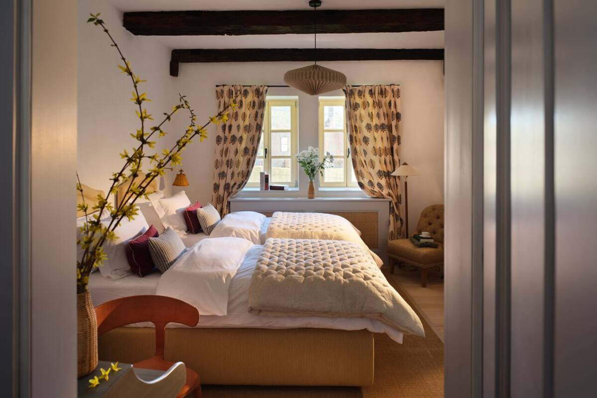 Quarto com uma cama grande, uma cadeira, um vaso com flores e uma janela com cortinas.