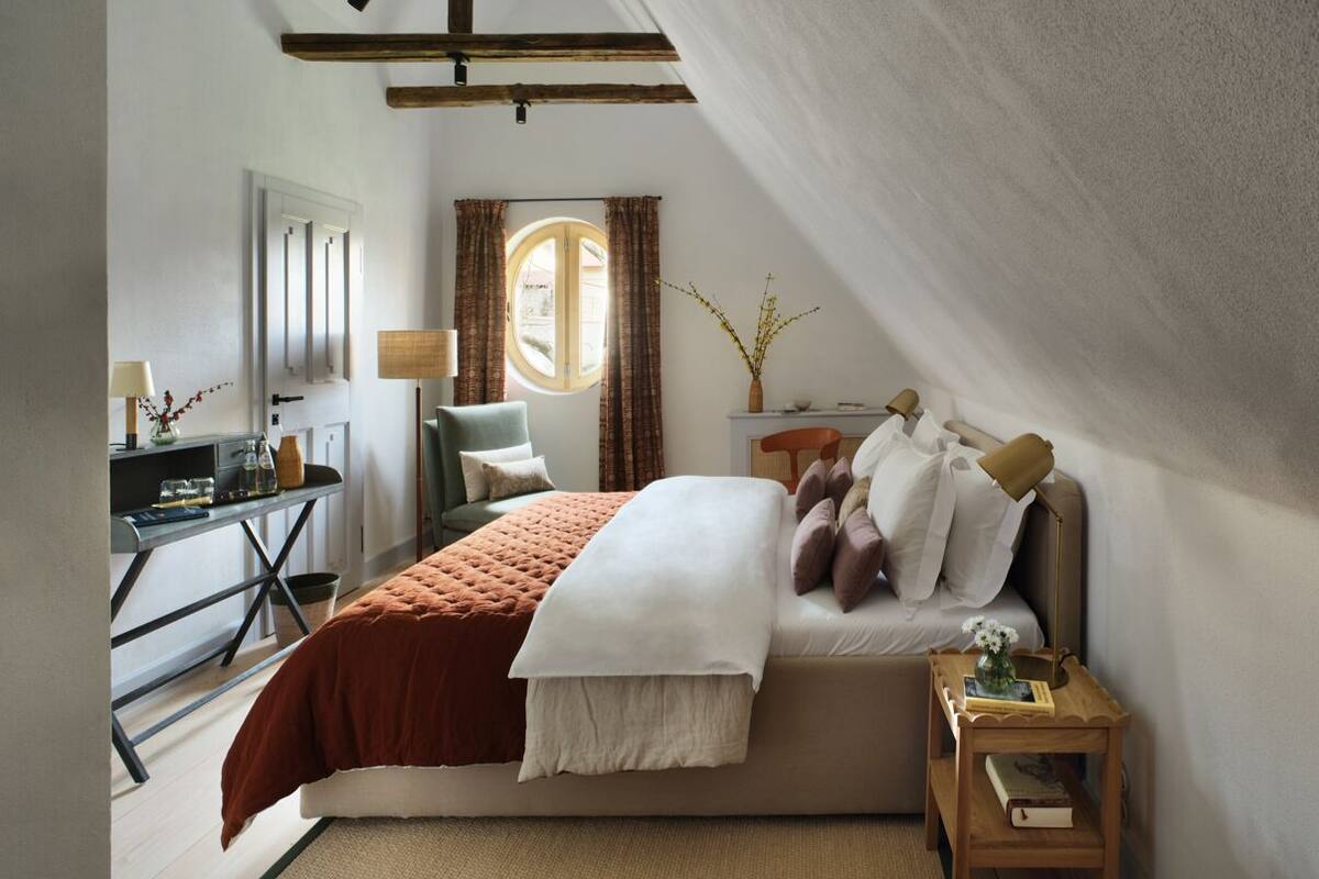 Quarto com cama, mesa de cabeceira, abajur e vista para o exterior pela janela.