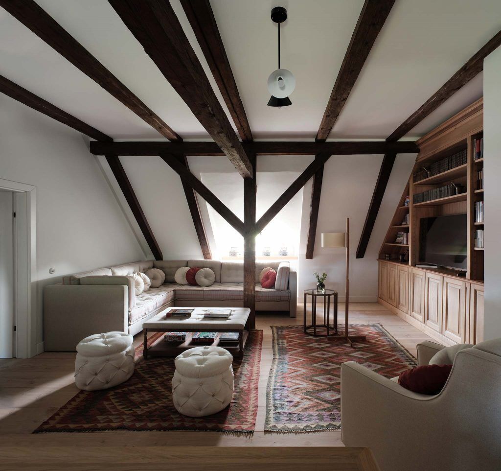 A loft-style living room with exposed wooden beams, a sofa set, ottomans, a rug, and a bookshelf with a television.