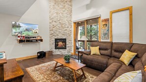 A cozy living room with a stone wall, a fireplace, a brown leather couch, and a large TV. - 2 Queen Beds | Rec Center Access | 3-Min Drive to Village | Near Lake & Trails (Truckee)