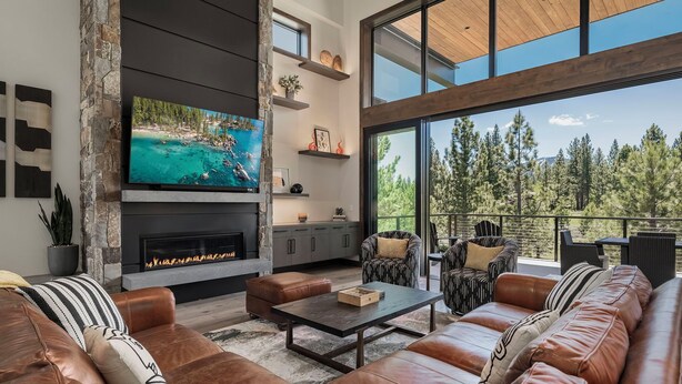 A modern living room with leather sofas, a flat-screen TV, a fireplace, and a large window overlooking a forested area. - Luxury Tahoe Estate | AC & Hot Tub | EV Charger| Golf Access, Near Lake & Trails (Truckee)