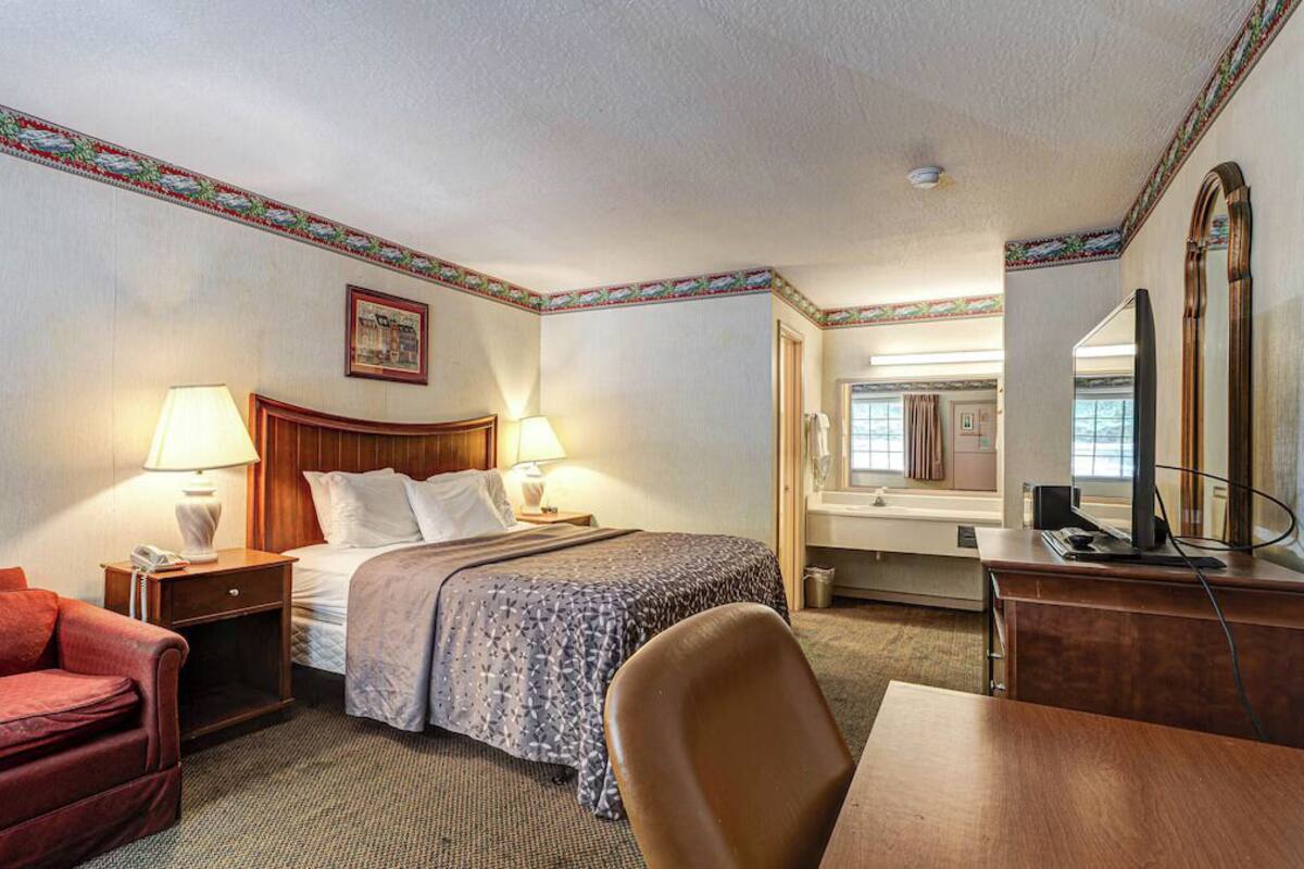 A hotel room with a bed, two bedside lamps, a red armchair, a wooden desk, and a large mirror.