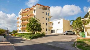 Un immeuble résidentiel à plusieurs étages, doté de balcons, d’un espace de stationnement et d’un palmier.