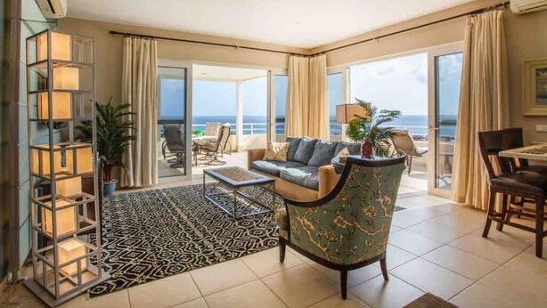 Un vaste espace de vie comprenant un canapé, des fauteuils, une table basse et un tapis à motifs. Une grande fenêtre offre une vue sur la mer.