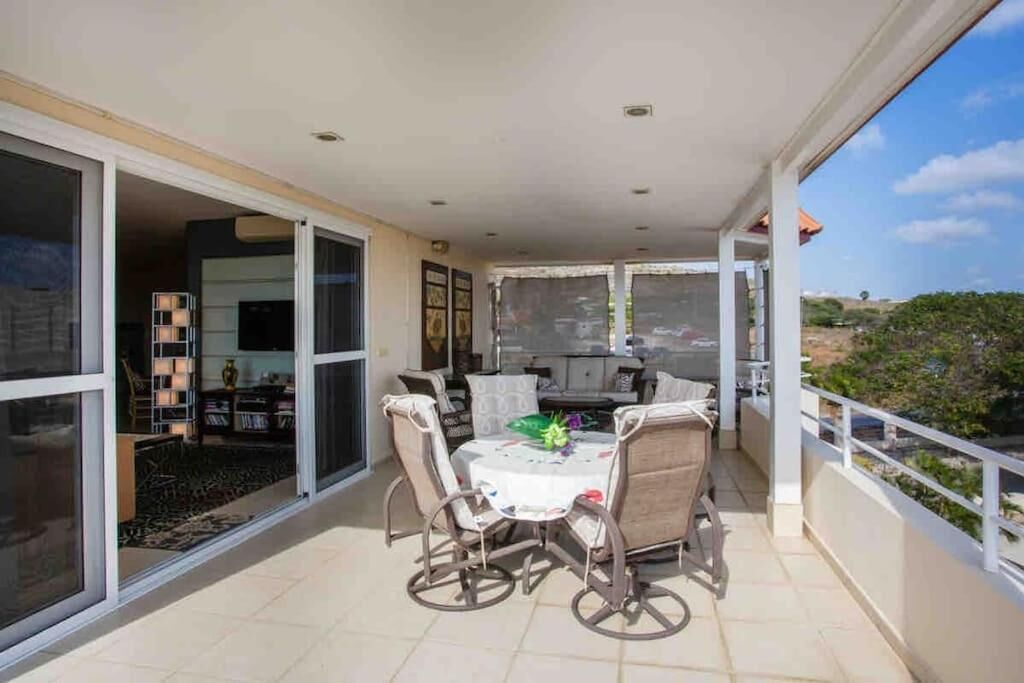 A balcony with a table and chairs, a sliding glass door leading to a living area, and a view of the landscape.