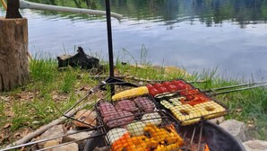 A campfire with various foods grilling, surrounded by a natural landscape and a body of water. - Tentrr-Riverfront Ritz (Lake Luzerne)