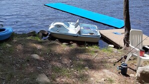 A lakeside setting with a boat docked on the shore, a blue paddleboard, and a white chair. - Tentrr-Riverfront Ritz (Lake Luzerne)