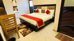 A hotel room with a bed, a wooden chair, a small trash bin, a ceiling fan, and a framed picture on the wall. - Hotel PSR Residency - near Temple & Ghat (Varanasi)