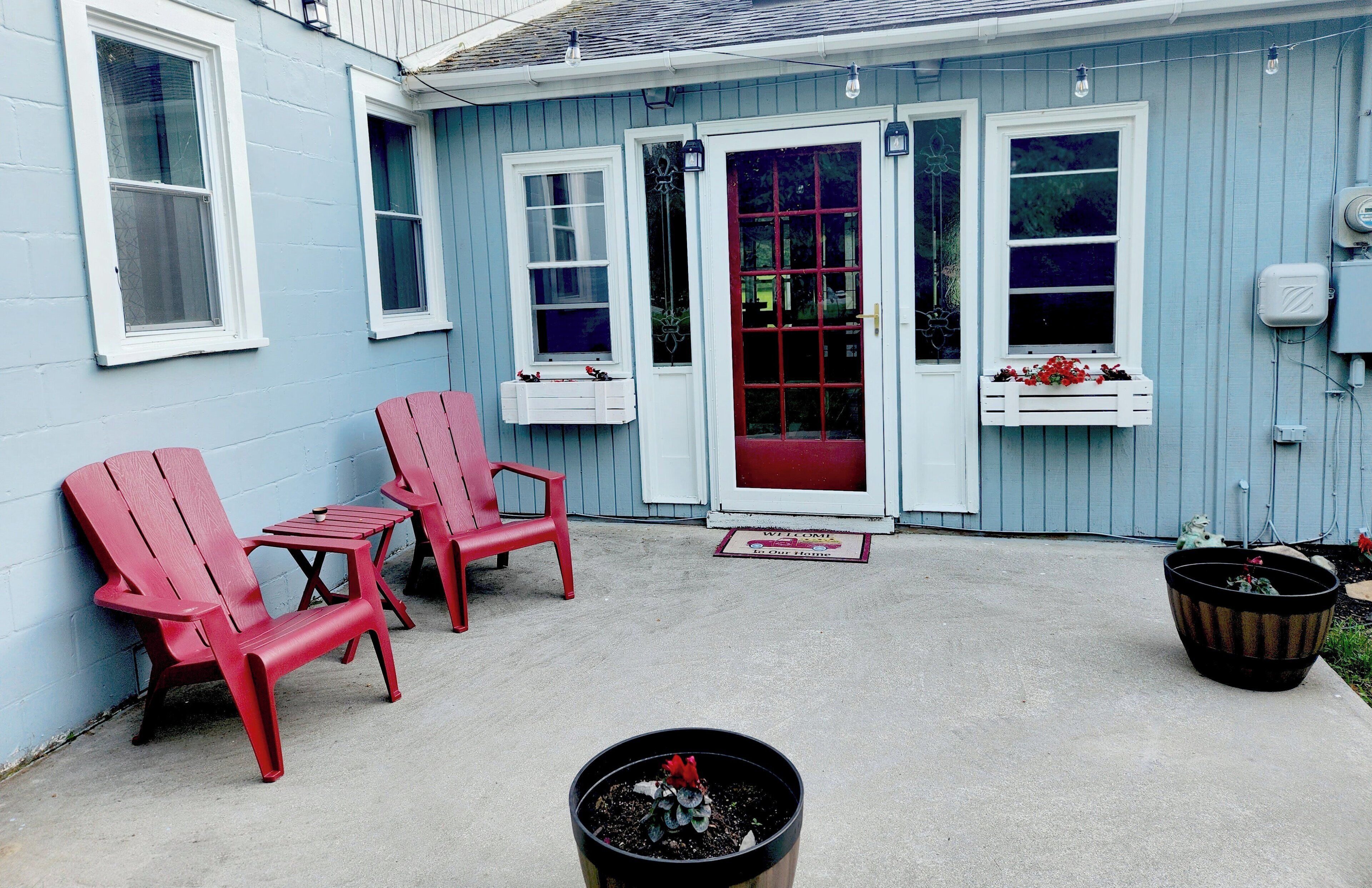 Une maison bleue avec une porte rouge, deux chaises rouges, une petite table et une plante en pot.