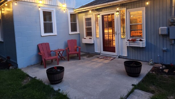 Een klein, laag gebouw met een overdekte patio, waar twee rode Adirondack-stoelen en een kleine tafel staan. Boven de patio hangen lichtslingers en er is een deur met een ruitpaneel die leidt naar de binnenruimte.