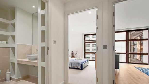 A modern bathroom with a white sink, shelves, and a mirror. A bedroom with a bed and a view of a building is visible through an open door. - 1 Bed South Quay Apartments Canary Wharf (London)