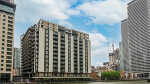 A modern high-rise building with multiple balconies, situated next to a river. - 1 Bed South Quay Apartments Canary Wharf (London)