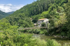 Una casa di pietra rustica con tetto in tegole rosse, un ponte di legno e un fiume in una valle verde e rigogliosa.