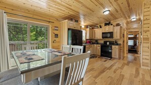 Een houten blokhut van binnen met een eettafel, stoelen, een keukenhoek met apparatuur, en uitzicht op het bos door grote ramen.