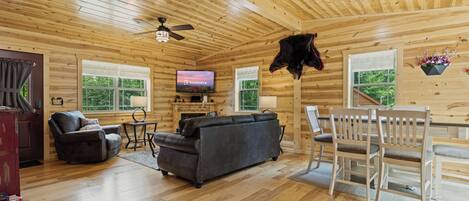 A wooden cabin interior with a sofa, chairs, a television, and a ceiling fan.