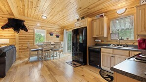 Een cabin interieur met houten wanden en vloeren, een eethoek met stoelen en een tafel, een keuken met een koelkast en wasbak, en een opgehangen dierenhoofd.