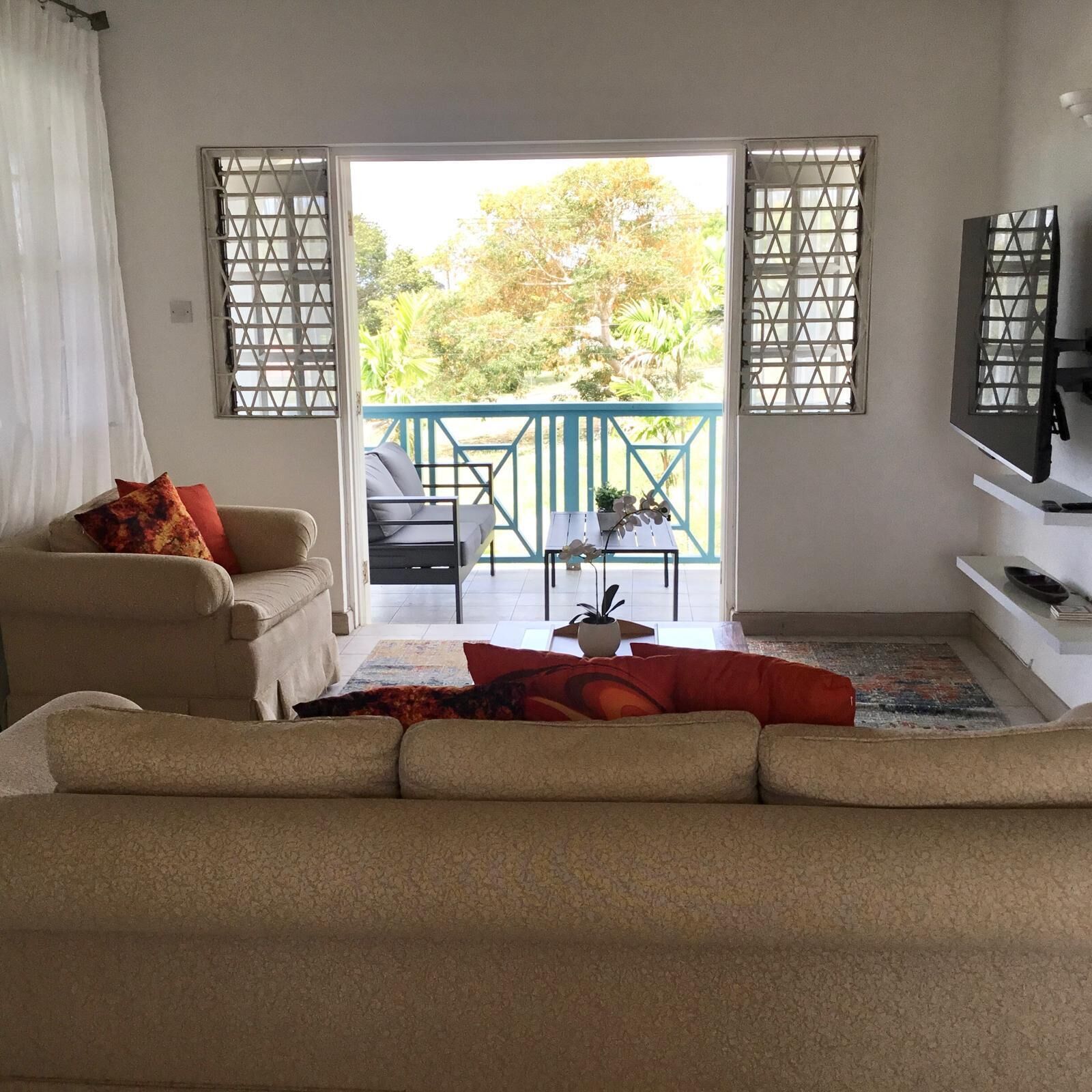 A living room with a sofa, a coffee table, and a TV on a stand. There is a balcony with a railing and a view of trees.