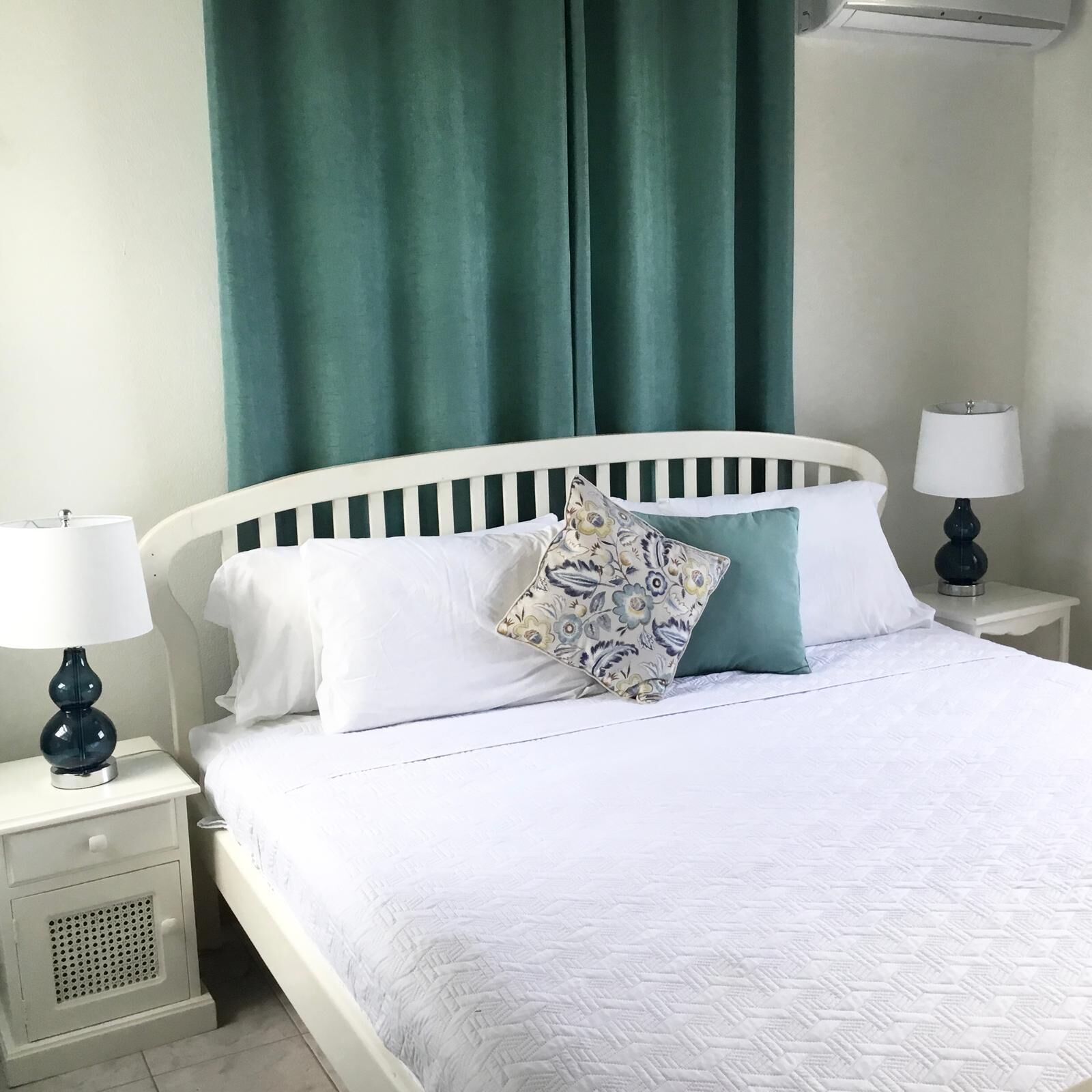 A neatly made bed with white bedding and a patterned pillow, flanked by two bedside tables with lamps, in a room with green curtains and a white headboard.