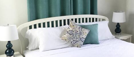 A neatly made bed with white bedding and a patterned pillow, flanked by two bedside tables with lamps, in a room with green curtains and a white headboard.