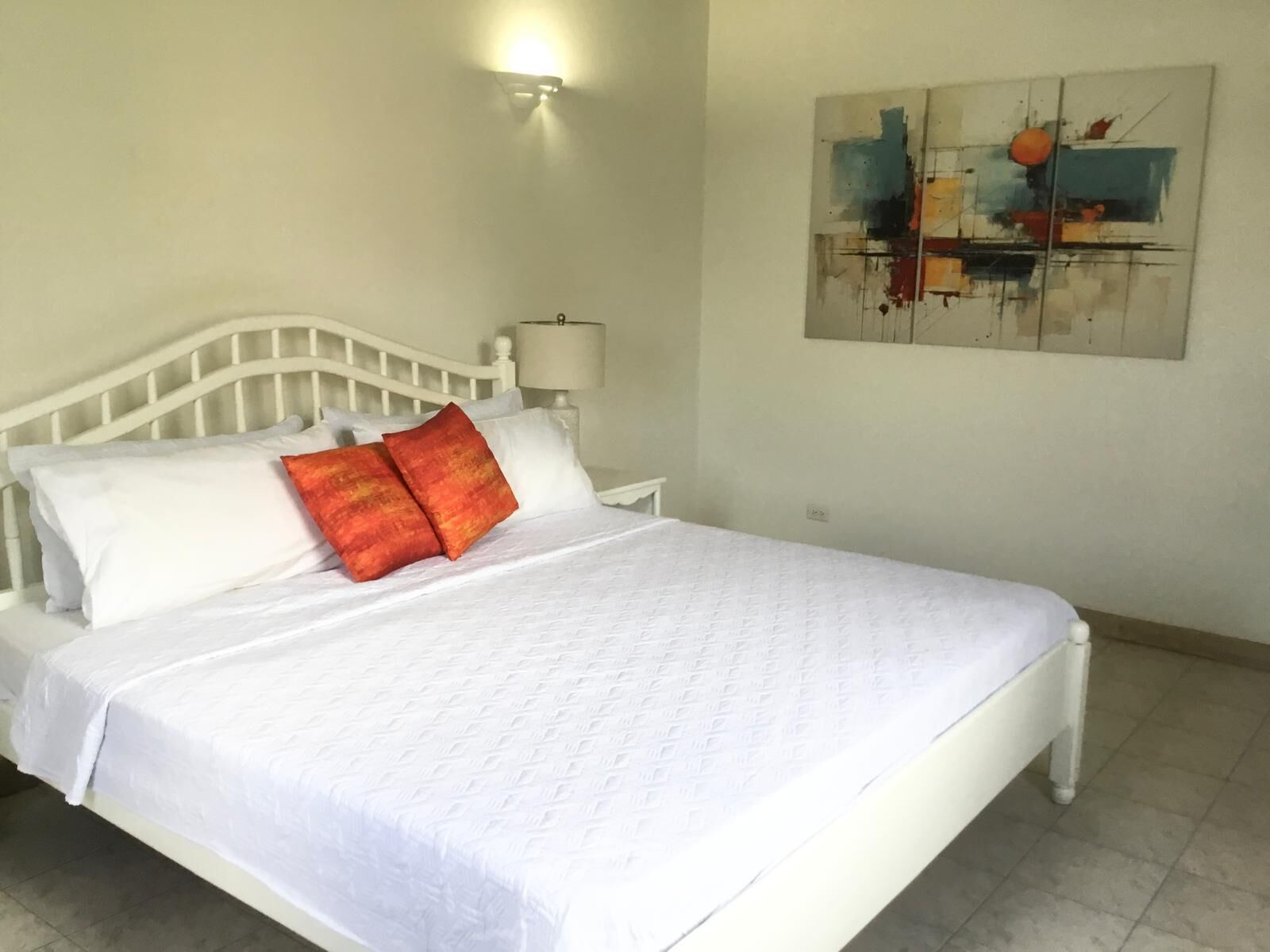 A neatly made bed with white bedding and two red pillows, a white nightstand with a lamp, and a painting on the wall.