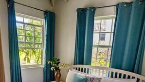 A bed with a white headboard and a patterned bedspread, a nightstand with a vase of flowers, and a window with blue curtains. - Delightful-3 bedroom with AC and Wifi near Oistins, St Lawrence Gap (Christ Church)