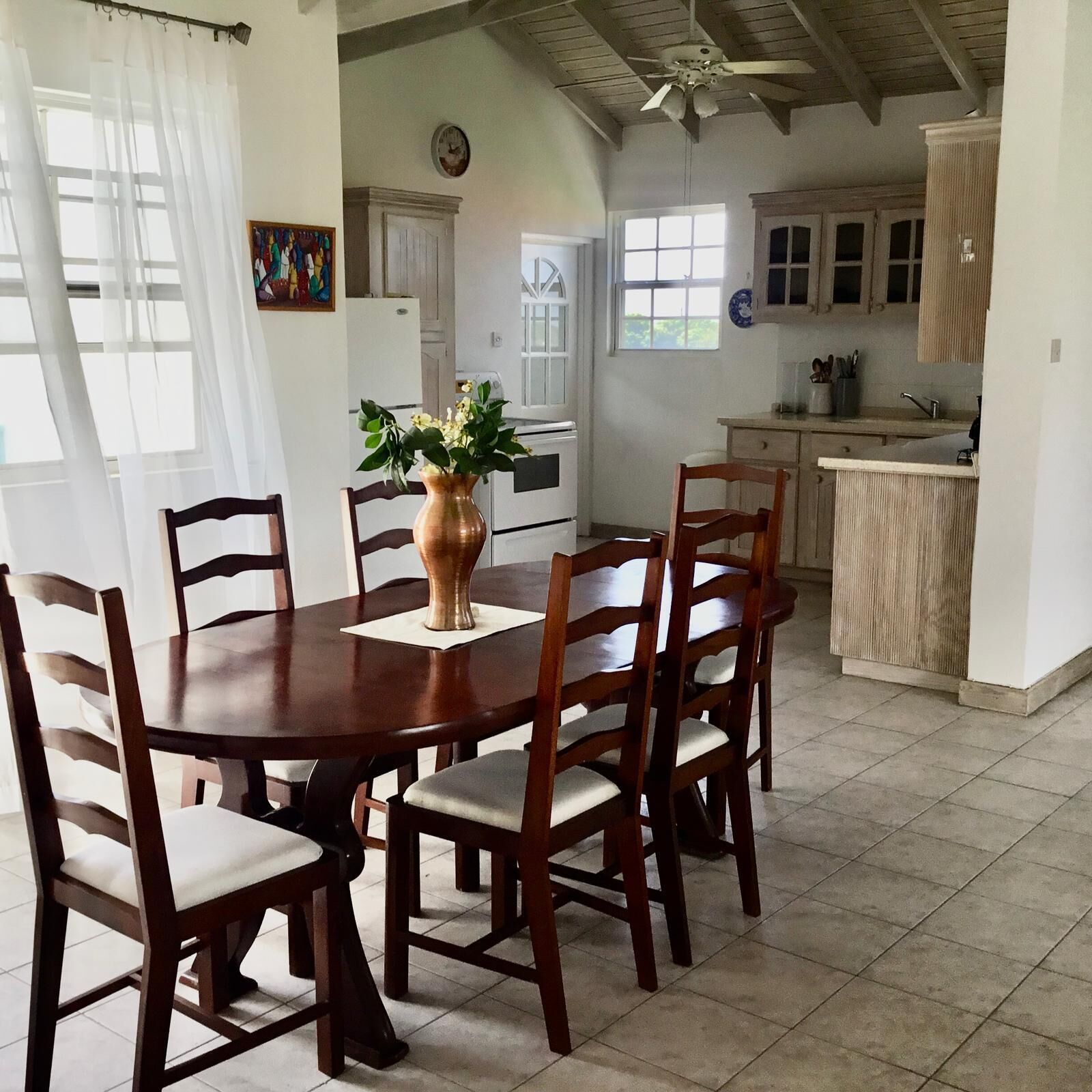 A dining area with a wooden table and chairs, a vase with flowers, and a kitchen in the background.
