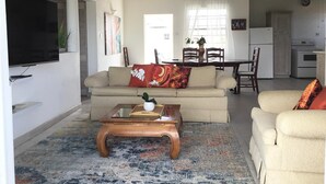 A living room with a sofa, coffee table, and TV. There is a dining area with a table and chairs. The room has a wooden ceiling and a kitchen in the background. - Delightful-3 bedroom with AC and Wifi near Oistins, St Lawrence Gap (Christ Church)