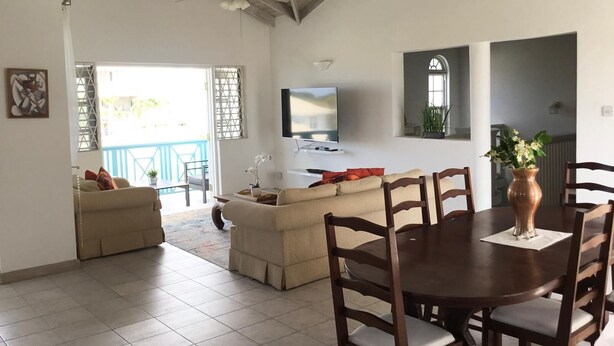 A spacious living area with a high ceiling, a TV mounted on the wall, and a dining table with chairs. - Delightful-3 bedroom with AC and Wifi near Oistins, St Lawrence Gap (Christ Church)