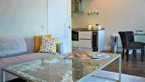 A modern living room with a glass coffee table, a sofa with patterned cushions, a dining area with a chair, and a kitchen in the background. - One Bed Penthouse Apartment in City Road (London)