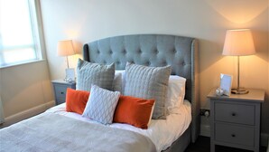 A neatly made bed with a grey headboard, two bedside tables with lamps, and a window with blinds. - One Bed Penthouse Apartment in City Road (London)