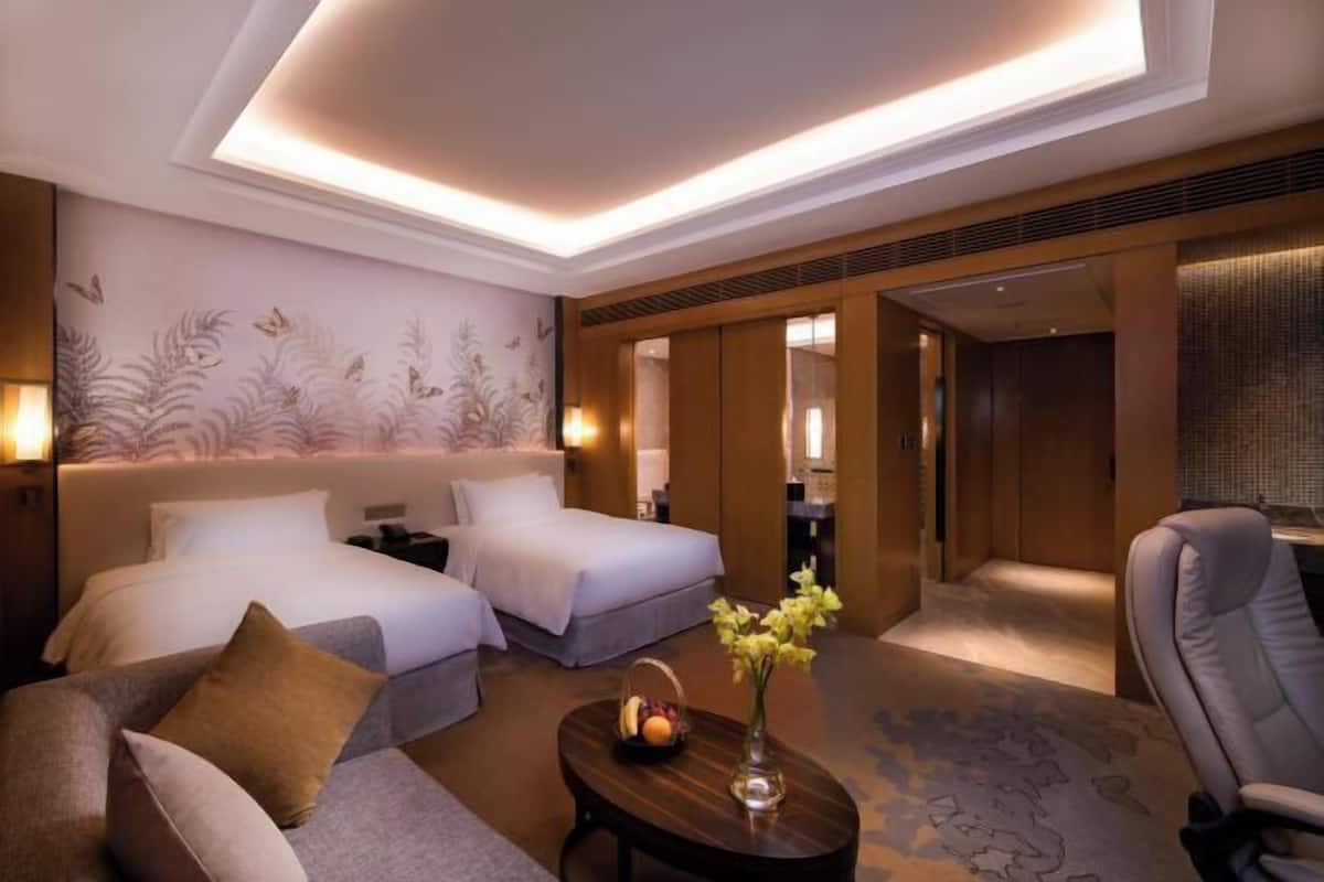 Ein Hotelzimmer mit zwei Betten, einem Schlafsofa, einem Couchtisch mit Obst und Blumen sowie Blick auf den Flur.