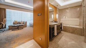 A modern hotel room with a large window, a sofa, a coffee table, and a bathroom with a bathtub and a large mirror. - Yongkang World Trade International Hotel (Yongkang)