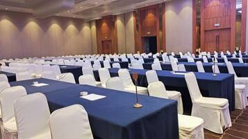 A conference room with rows of tables and chairs, a stage at the front, and a large window on the right.