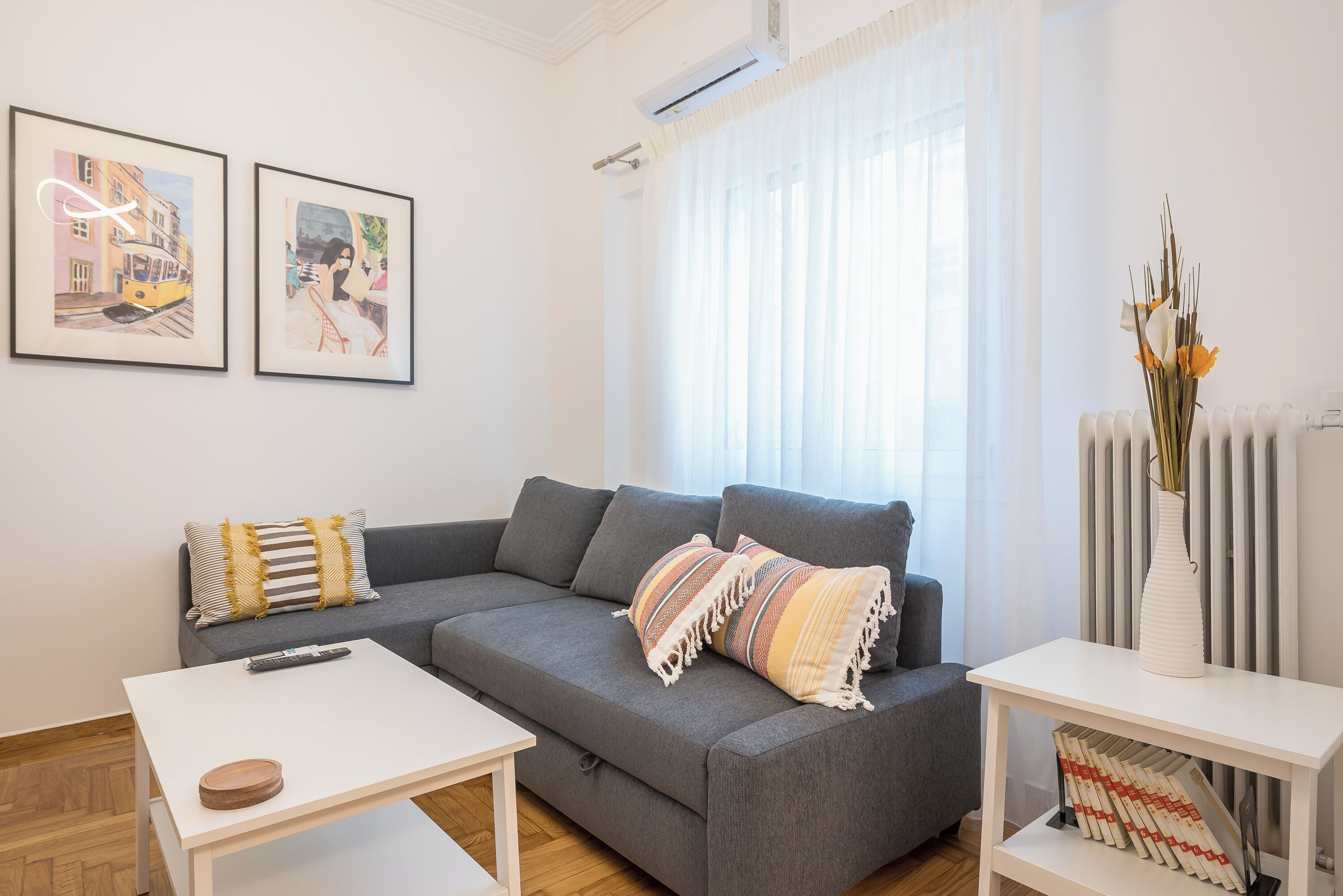 En moderne stue med en grå sofa, et hvidt sofabord og et hvidt konsolbord med en vase. To indrammede kunstværker hænger på væggen, og der er en radiator til højre.