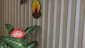 En stribet væg med en træstol, en potteplante og dekorative udskæringer, heriblandt en tegneseriefigur, en ild og et skilt med teksten "Cadê a BARRACA do Beijo?".