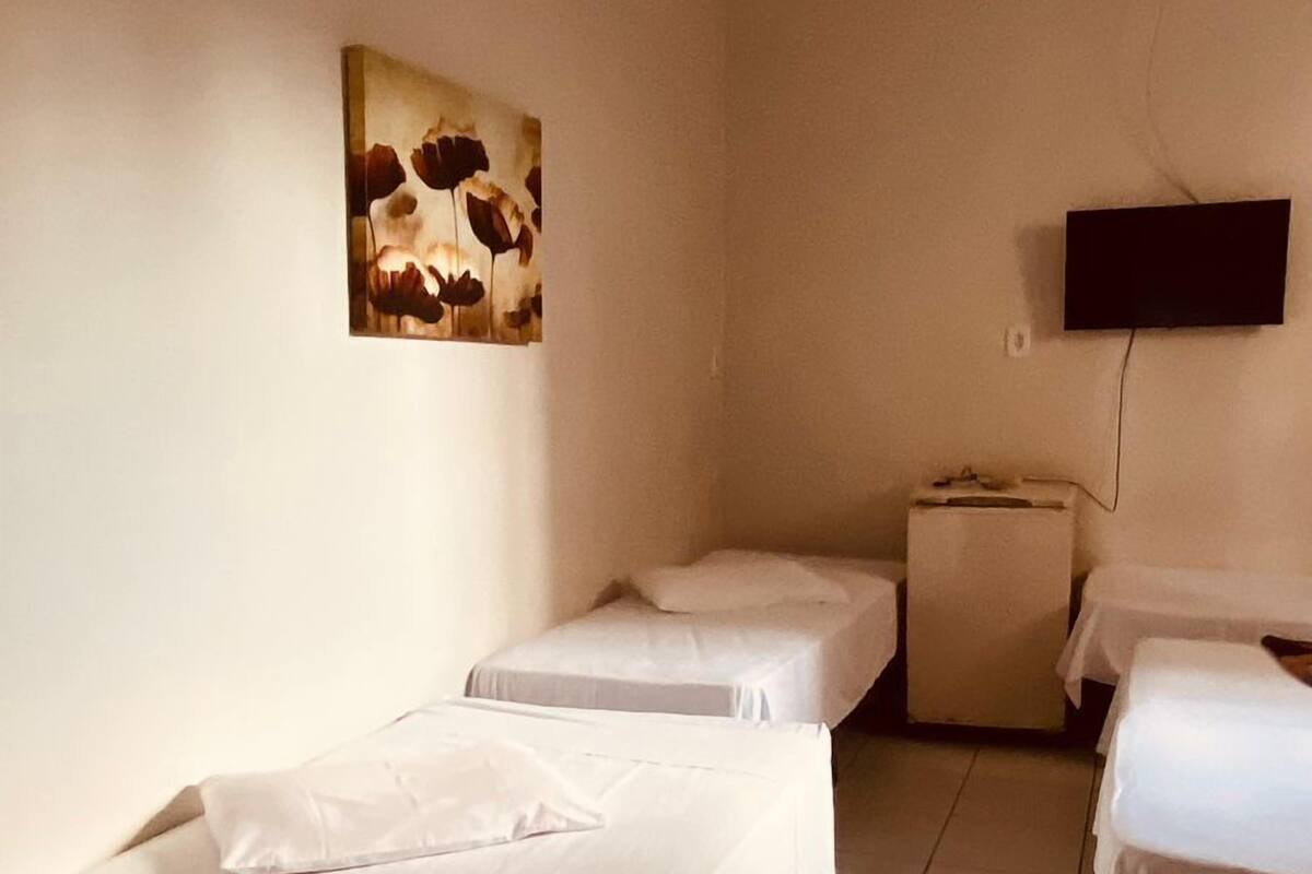 Ein Hotelzimmer mit drei Einzelbetten, einem kleinen Tisch und einem an der Wand befestigten Fernseher.