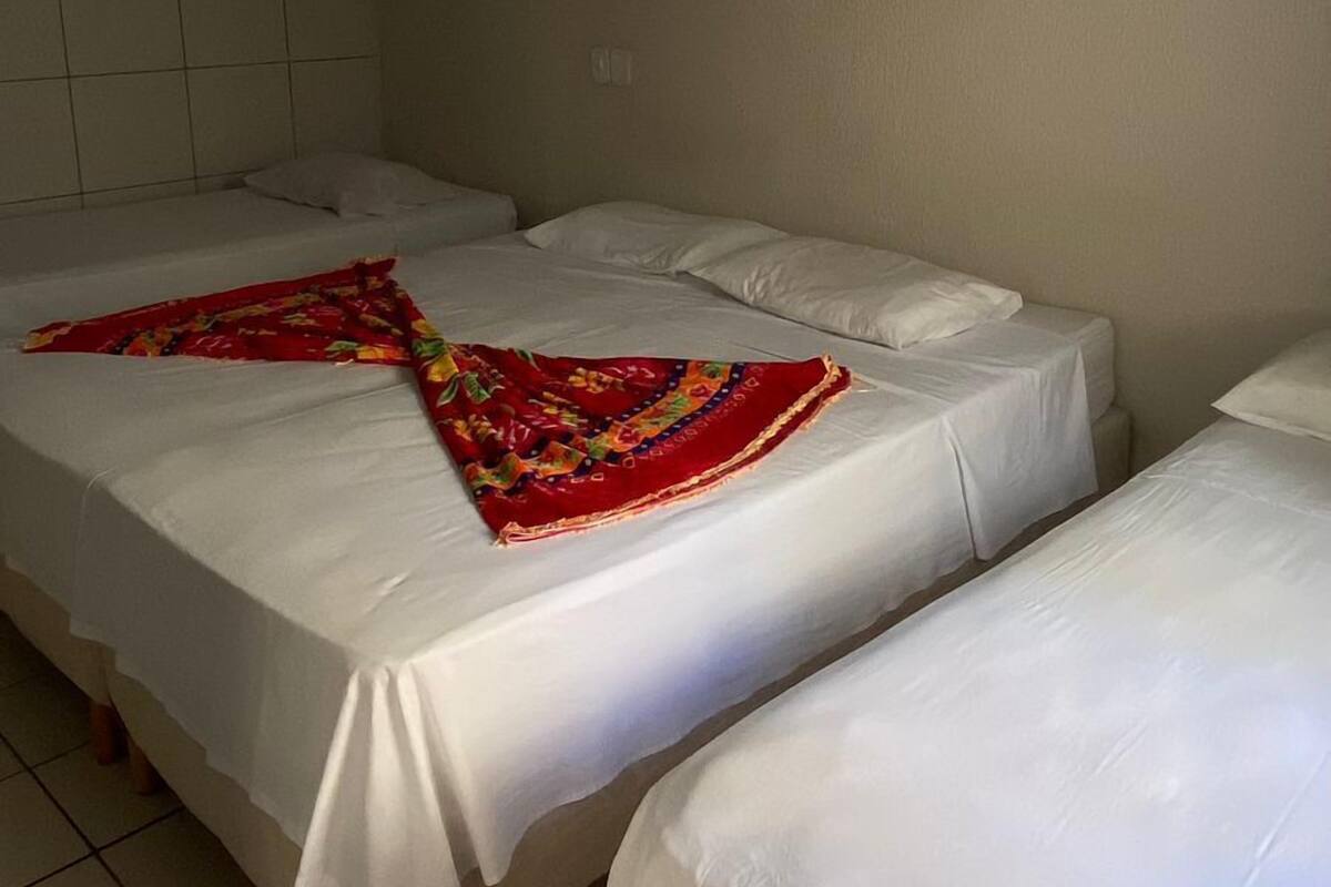 Ein Zimmer mit drei Einzelbetten, jedes mit weißer Bettdecke und bunter Decke. Rechts befindet sich ein Wandhaken.