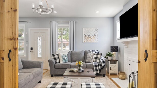 A living room with a grey sofa, a coffee table, and two ottomans. There is a TV mounted on the wall and a window with curtains. - The Bell House - Contemporary Comfort Near Wards Road with a Luxurious Hot Tub! (Lynchburg)