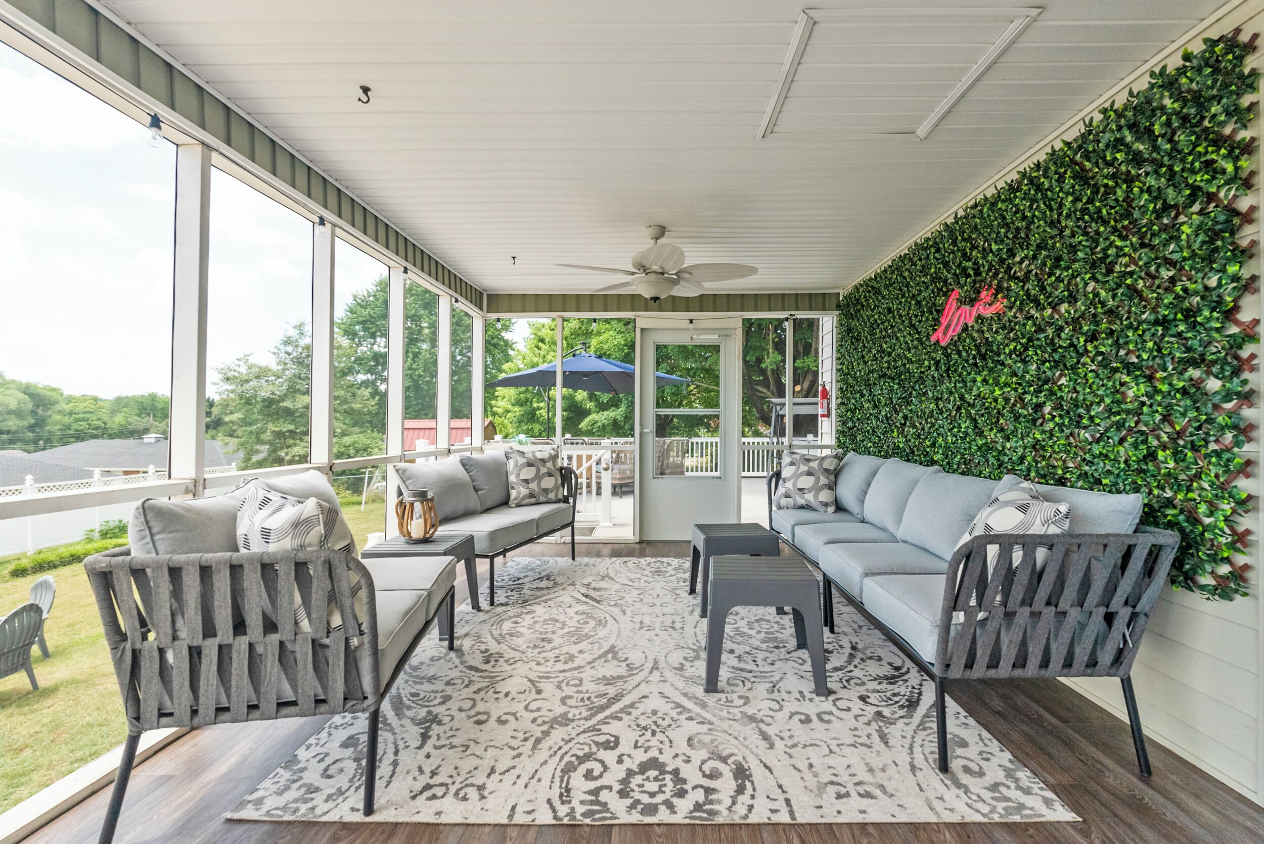 Une terrasse couverte avec un ventilateur de plafond, un canapé gris, un tapis à motifs et un mur vert orné d’un panneau indiquant le mot « Amour ».