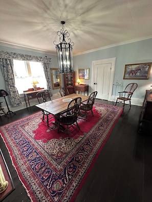A dining room with a large wooden table, six chairs, a chandelier, a patterned rug, and a window with curtains.