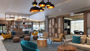 Um saguão moderno de hotel com um sofá, cadeiras e uma mesa de centro. Há luminárias pendentes e uma grande janela ao fundo.