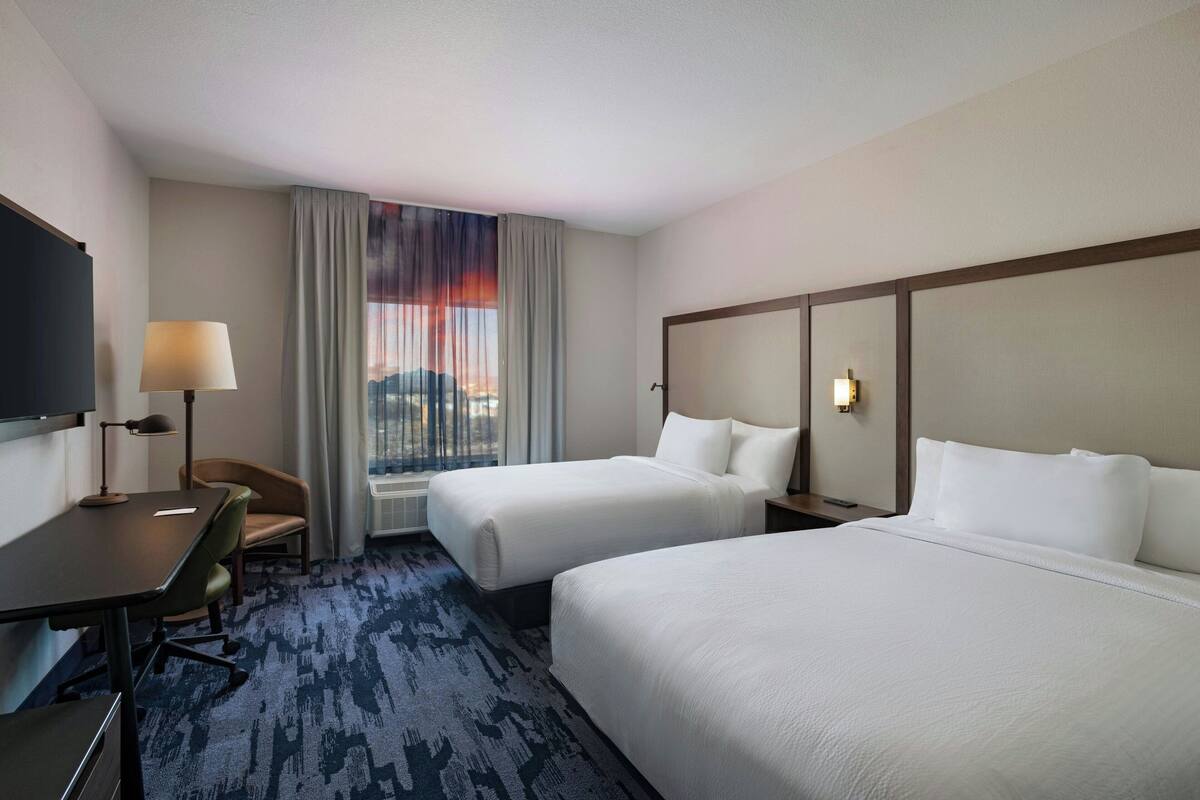 A hotel room with two beds, a desk, a chair, a TV, and a window with curtains.