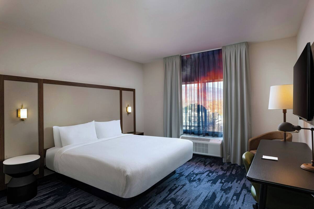 A hotel room with a large bed, a desk with a lamp, a chair, and a window with curtains.
